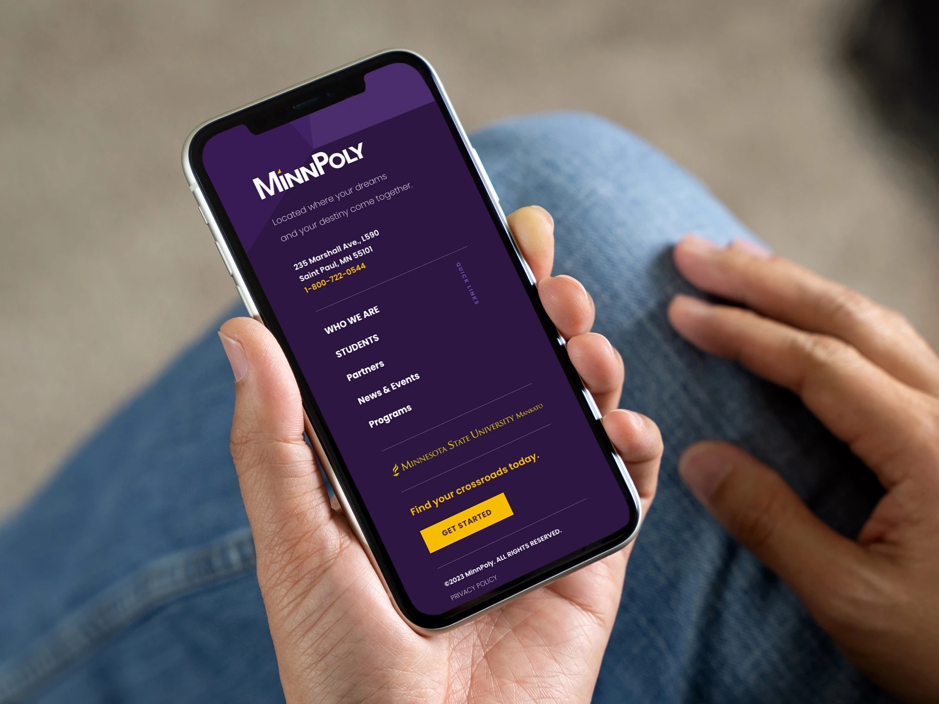 Person holding a smartphone displaying the MinnPoly website footer, showcasing the mobile-responsive design developed by LXE Digital with navigation, contact details, and a call-to-action